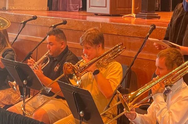 Three musicians playing brass instruments, seated in a row on stage, with music stands in front of them.